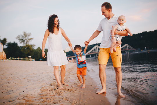 Concept Family Beach Vacation. Family Of Four People Mom, Dad And Brothers Children Boys Walking, Want Barefoot On The River Bank By The Hand At Sunset In The Summer