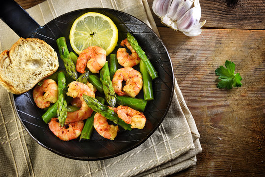 fried prawns or shrimps with  green asparagus and a lemon slice in a black iron pan, garlic, bread and parsley on a rustic wooden table, top view from above, copy space