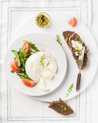 Sliced Italian cheese Burrata (mozzarella) with fresh arugula, tomatoes, olive oil, sauce pesto and whole grain bread toasts on a white plate and a wooden light background. Flat lay, top view