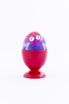 Pink and purple painted Easter egg with funny cartoon style face in a red plastic egg cup and white background