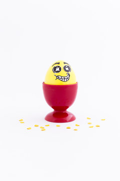 Yellow painted Easter egg with funny cartoon style face in a red plastic egg cup and yellow confetti on white background