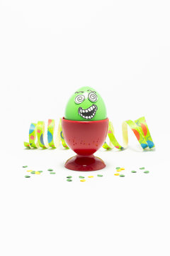 Green painted Easter egg with funny cartoon style face in a red plastic egg cup, colorful paper streamer and confetti on white background