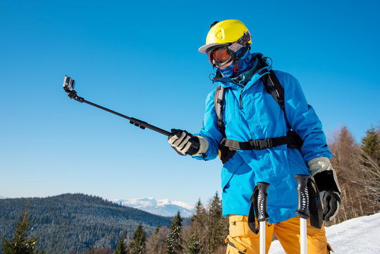 Shot of a skier in winter clothing taking a selfie using monopod for his camera. Blue sky and winter forest on the background skiing activity sports concept