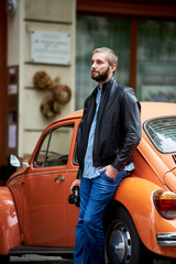 Profile of a man with the beard leaning on orange retro car and holding in hand a professional photo camera against the backdrop of urban architecture in Budapest. Close-up