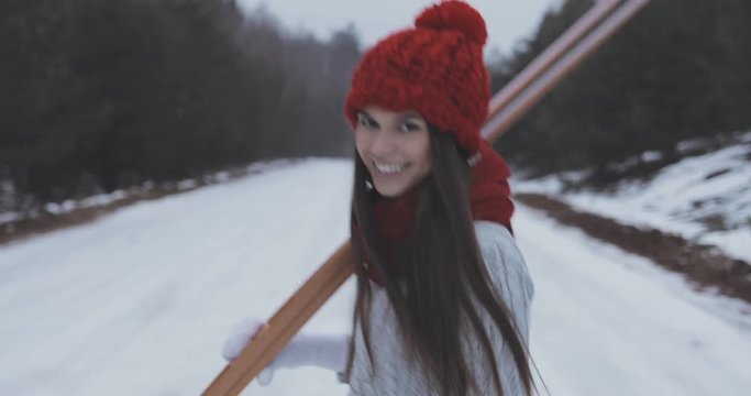 attractive sporty brunnete walking, turning back and smiling, carrying ski (skis), wering grey sweater and red scarf and cap. winter snowy forest as background