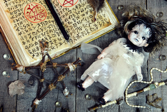Scary doll, pentagram and open magic book with evil symbols on witch table. Occult, esoteric, divination and wicca concep. No foreign text, all symbols on pages are fantasy, imaginary ones