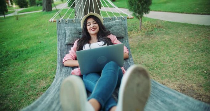 young successful girl blogger lies in a hammock with a laptop in the park, enjoys life at remote work