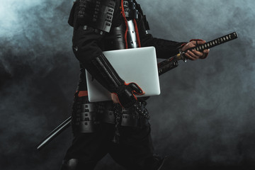 cropped shot of samurai in traditional armor with laptop taking out sword on dark background with...