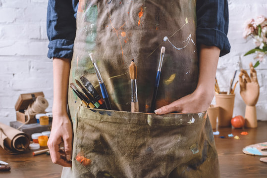 cropped image of artist with brushes and hand in apron pocket