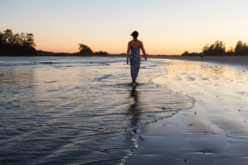 Woman in a casual dress walking on a sandy beach during a vibrant sunset. Taken at Chesterman, Tofino, Vancouver Island, British Columbia, Canada.
