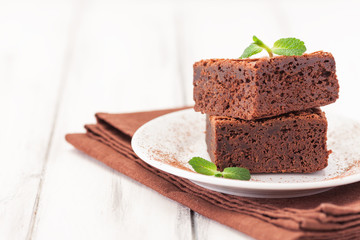 Chocolate brownie square pieces in stack on white plate decorated with mint leaves and cocoa powder on white vintage wooden background. American traditional delicious dessert. Close up photography