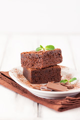 Chocolate brownie square pieces in stack on white plate decorated with mint leaves and cocoa powder on white vintage wooden background. American traditional delicious dessert. Close up photography