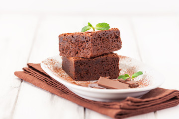 Chocolate brownie square pieces in stack on white plate decorated with mint leaves and cocoa powder on white vintage wooden background. American traditional delicious dessert. Close up photography