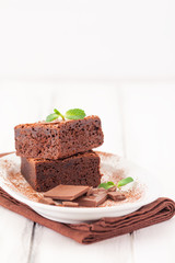 Chocolate brownie square pieces in stack on white plate decorated with mint leaves and cocoa powder on white vintage wooden background. American traditional delicious dessert. Close up photography