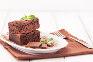 Chocolate brownie square pieces in stack on white plate decorated with mint leaves and cocoa powder on white vintage wooden background. American traditional delicious dessert. Close up photography