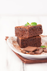Chocolate brownie square pieces in stack on white plate decorated with mint leaves and cocoa powder on white vintage wooden background. American traditional delicious dessert. Close up photography