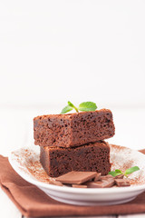 Chocolate brownie square pieces in stack on white plate decorated with mint leaves and cocoa powder on white vintage wooden background. American traditional delicious dessert. Close up photography