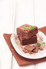 Chocolate brownie square pieces in stack on white plate decorated with mint leaves and cocoa powder on white vintage wooden background. American traditional delicious dessert. Close up photography