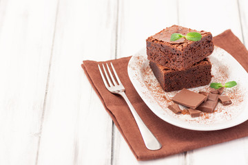 Chocolate brownie square pieces in stack on white plate decorated with mint leaves and cocoa powder on white vintage wooden background. American traditional delicious dessert. Close up photography