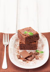 Chocolate brownie square pieces in stack on white plate decorated with mint leaves and cocoa powder on white vintage wooden background. American traditional delicious dessert. Close up photography