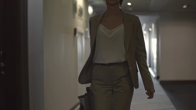 Elegant young businesswoman with short haircut and eyeglasses walking through corridor in hotel and pulling her luggage