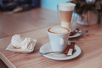 Cup of coffee, a transparent glass of latte and two hand made marshmellows on a light wooden table surface, nice turquoise colored wall in a blurred background.