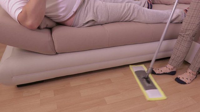 Lazy man with beer on sofa near woman who cleaning floor
