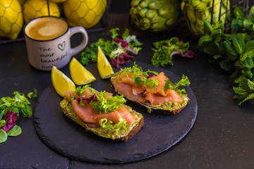 morning breakfast with hot coffee and a sandwich with avocado, soft cheese, lemon and red fish trout on a dark background