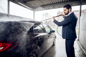 Side view of young handsome serious stylish bearded businessman in the suit cleaning the car with a water gun in the manual self-service washing station.