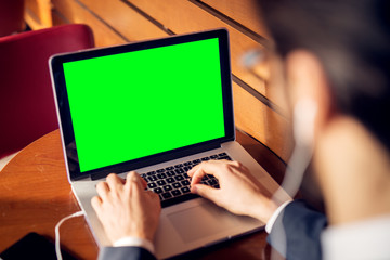 Fototapeta na wymiar Rear close up focus view laptop with blank green screen and hands of young successful stylish focused businessman in the suit with earphones typing in a cafe or restaurant near the wooden wall.