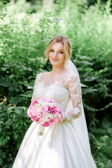 Bride in classy dress with long sleeves holds pink wedding bouquet standing in the park