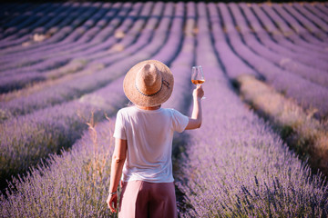 Fototapeta premium Młoda kobieta pije wino różane w polu lawendy słońca, stojąc z powrotem do kamery, Provence, południowej Francji