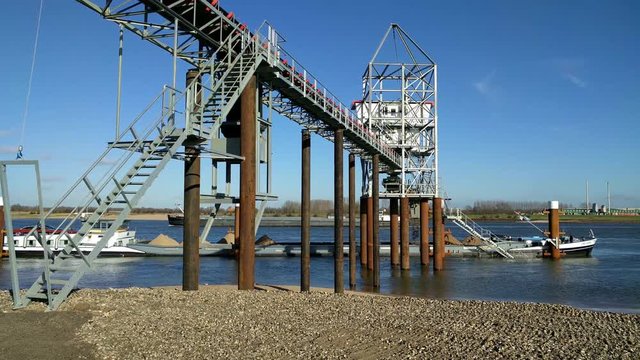 Loading a barge with sand using a conveyor + inland ship sailing downstream in background.