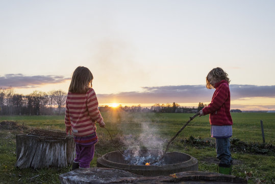 Two young girls digging in fire with sticks at sunset