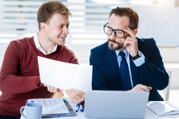 Funny thing. Appealing pleased two male colleagues emerging at work while  examining document and man touching glasses 