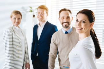 Sincere smile.  Adorable brunette charming female  colleague smiling while staring at the camera and posing on the blurred background 