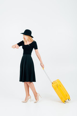 A beautifully dressed girl in a dark dress and a big hat looks at a clock with a yellow suitcase.