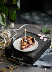 Fototapeta na wymiar A piece of a birthday cherry cake with a burning one white candle. Atmospheric beautiful food birthday photo with home cherry pie, flowers, ribbons and decoration.
