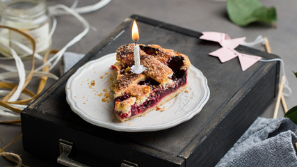 A piece of a birthday cherry cake with a burning one white candle. Atmospheric beautiful food birthday photo with home cherry pie, flowers, ribbons and decoration.