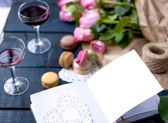 Obraz na płótnie Canvas A bouquet of roses is pink, in gray paper and a white heart. Open notebook. Two glasses of red wine. Sweet pasta macaroons of different colors, on Valentine's Day. A free place for text or advertising