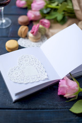 A bouquet of roses is pink, in gray paper and a white heart. Open notebook. Two glasses of red wine. Sweet pasta macaroons of different colors, on Valentine's Day. A free place for text or advertising
