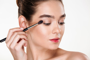 Fototapeta premium Close up portrait of elegant woman with clean shining skin applying eye shadow using brush isolated over white wall