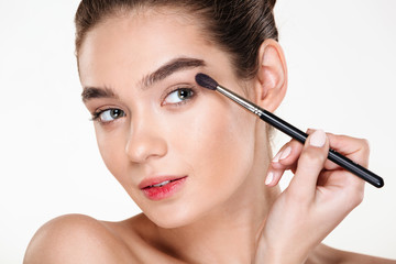 Fototapeta premium Close up portrait of sensual woman with clean shining skin applying eye shadow using brush isolated over white background