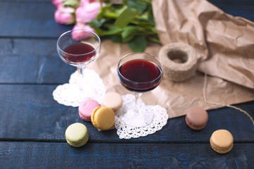 Two glasses of red wine on a dark table, a bouquet of pink roses in brown paper. Two white hearts and macaroni biscuits. Free space for text or postcards.