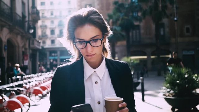 Close-up of attractive young businesswoman walking on urban street and using modern smartphone outside, sunshine, slow motion