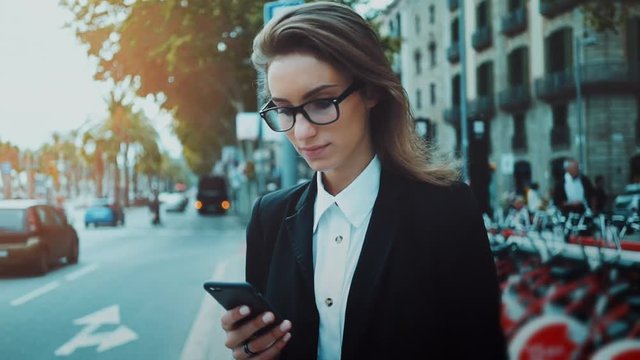 Close-up portrait of young beautiful businesswoman wearing eyeglasses and suit using her smartphone while standing on urban street, slow motion