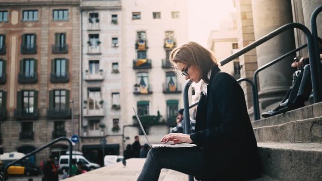 Close-up of young attractive businesswoman using laptop on a break outdoors, beautiful female student sitting on the stairs of University, slow motion