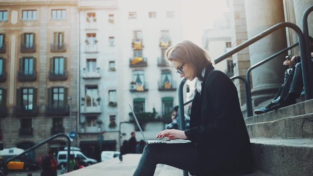 Side view of young hipster girl sitting on stairs and using laptop, attractive businesswoman typing on portable computer keyboard outdoors, slow motion