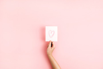 Obraz premium Valentine's Day composition. Female hand holding card with heart symbol on pale pink background. Flat lay, top view Love concept.