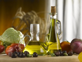 olive oil glass cruets, scattered olives and vegetables. front view on kitchen table. wooden wall and white curtain are in the background.